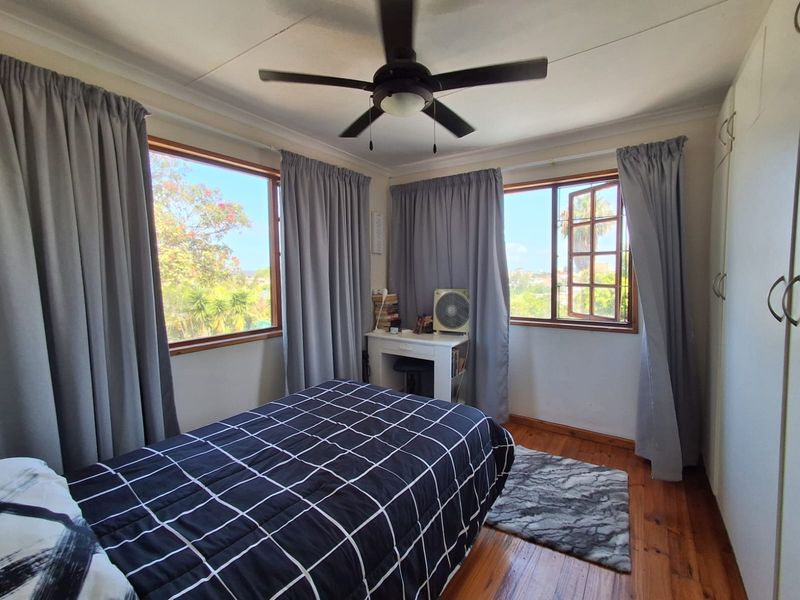 A bright bedroom featuring polished timber flooring, built-in cupboards, a ceiling fan, and large windows providing natural light and garden views.
