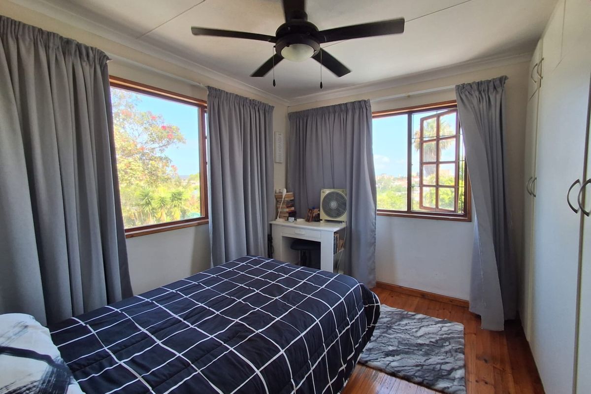 A bright bedroom featuring polished timber flooring, built-in cupboards, a ceiling fan, and large windows providing natural light and garden views.
