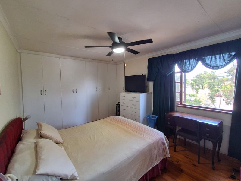 A bedroom featuring timber flooring, built-in cupboards, and a ceiling fan, with natural light provided by a large window.
