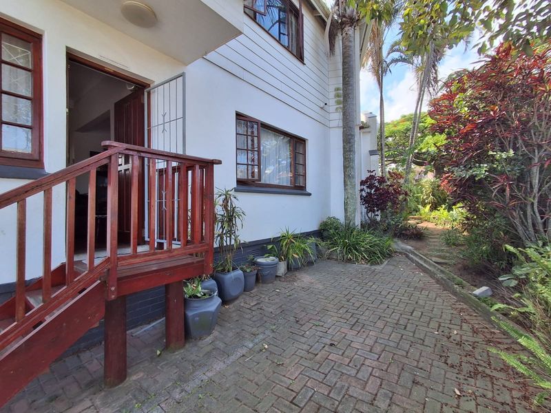 Rear exterior view of the property featuring a timber staircase leading to the entrance, paved patio area, and established garden surroundings.