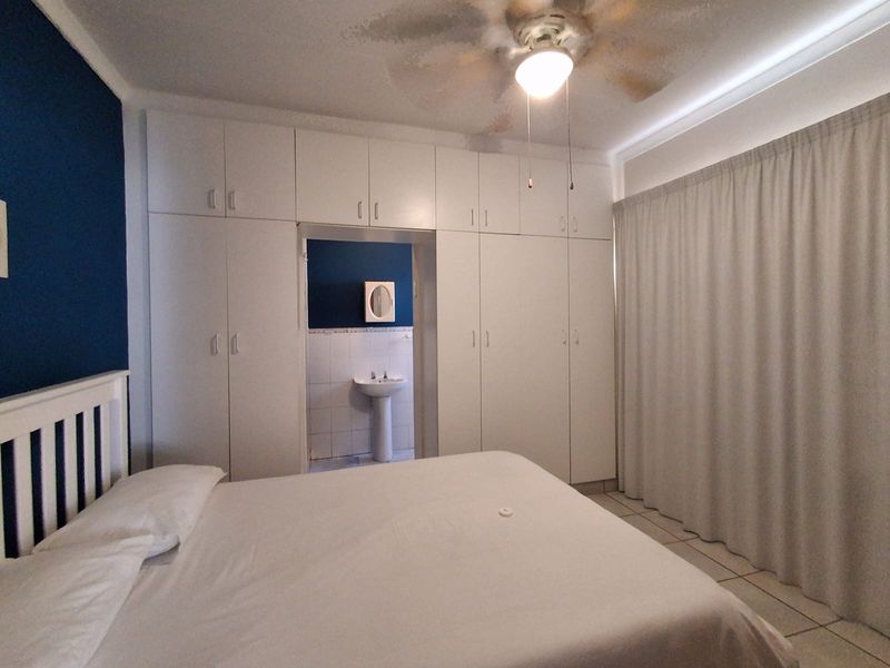 Bedroom featuring extensive built-in cupboards, a ceiling fan, and an en-suite area with a pedestal basin. Requires ceiling repairs.