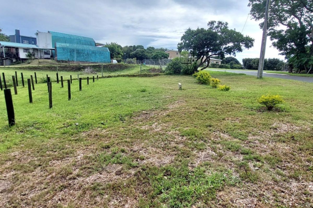 A view of a vacant residential plot featuring a grass lawn, boundary posts, and mature trees, situated along a street.