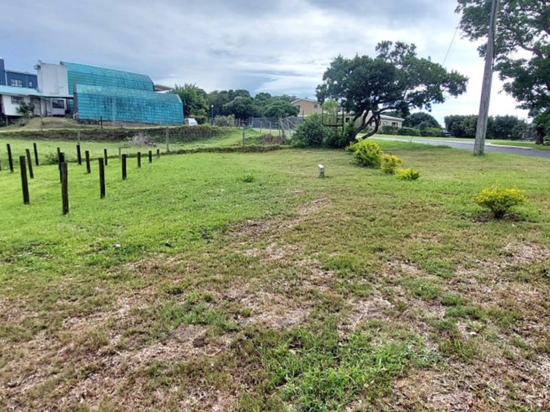 A view of a vacant residential plot featuring a grass lawn, boundary posts, and mature trees, situated along a street.