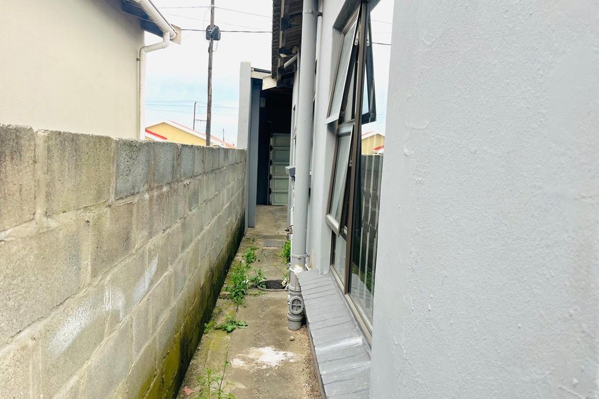 A narrow side passage between the house and a concrete boundary wall, featuring aluminium windows and a concrete walkway with some vegetation growth.