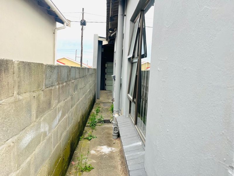 A narrow side passage between the house and a concrete boundary wall, featuring aluminium windows and a concrete walkway with some vegetation growth.