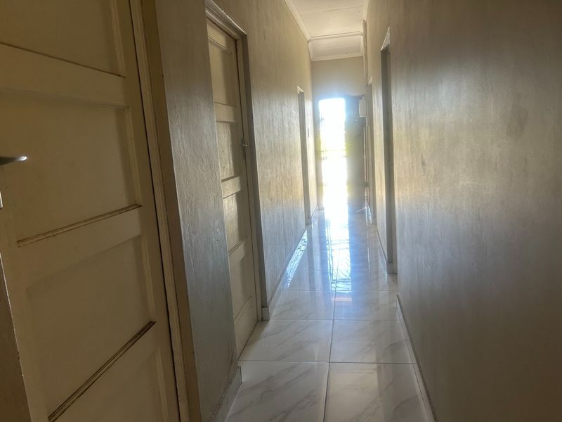 A bright, empty hallway featuring tiled flooring and multiple internal doors, leading towards a sunlit exit.
