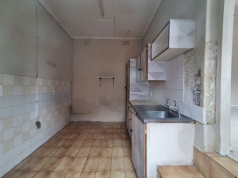 A small, dated kitchen area featuring a stainless steel sink, tiled splashback, and basic cabinetry, currently in need of renovation and cosmetic repairs.