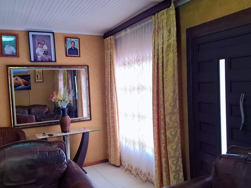 A living room featuring tiled flooring, a timber panelled ceiling, and a solid panelled entrance door with natural light filtering through window curtains.