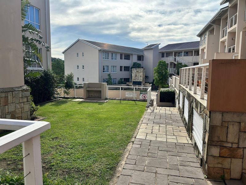 A well-maintained communal garden area featuring a paved walkway, built-in braai facilities, and stone-clad architectural details within the apartment complex.