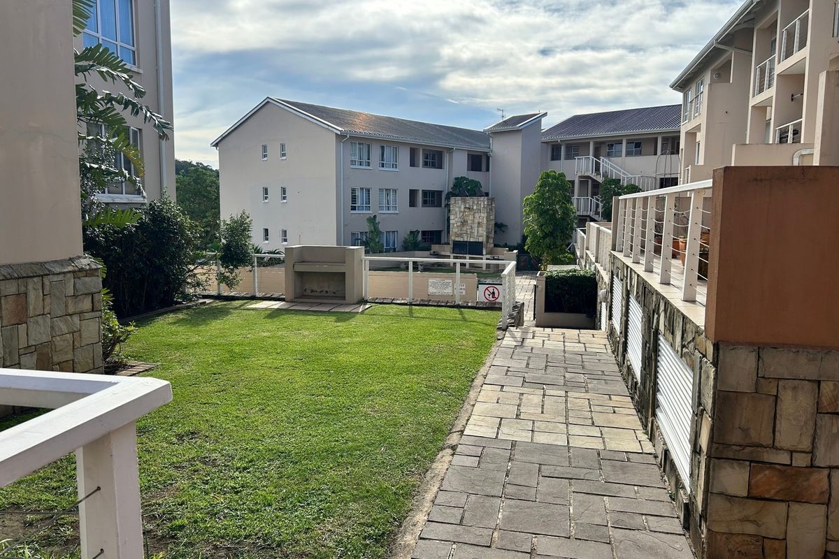 A well-maintained communal garden area featuring a paved walkway, built-in braai facilities, and stone-clad architectural details within the apartment complex.