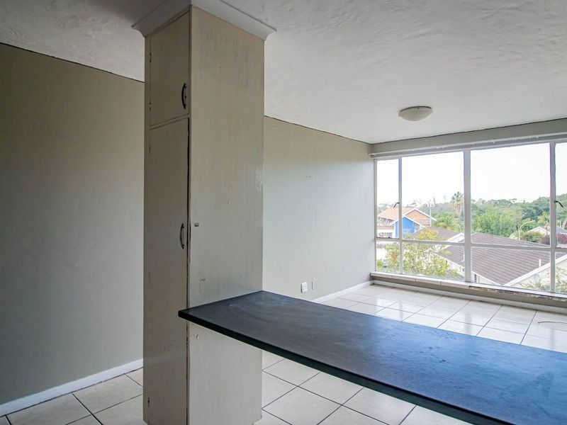 An empty open-plan living area featuring tiled floors, a built-in breakfast bar, and large windows providing natural light and neighborhood views.