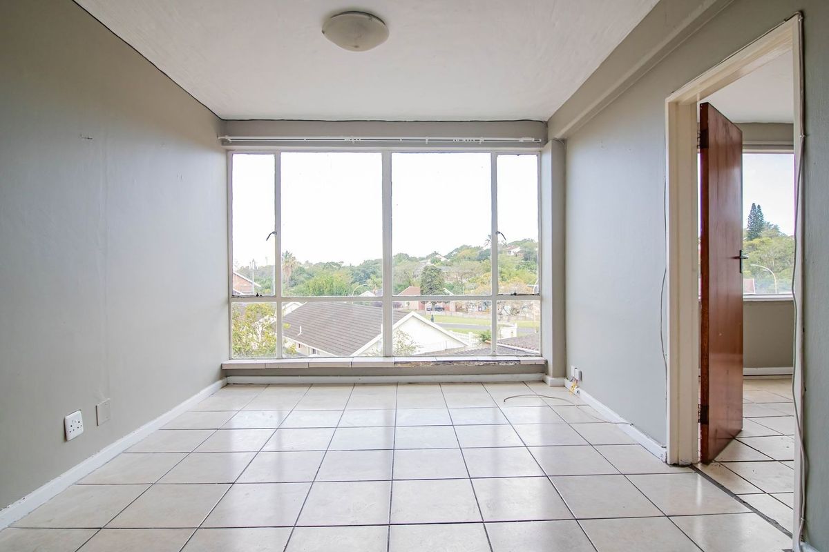 An empty living room featuring tiled flooring, a large window with natural light, and a view of the surrounding neighborhood.