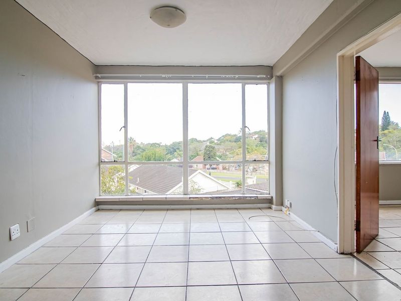 An empty living room featuring tiled flooring, a large window with natural light, and a view of the surrounding neighborhood.