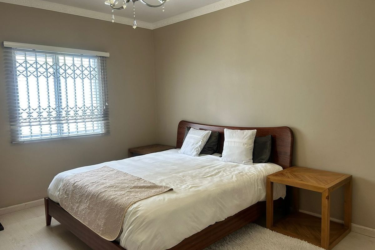 A well-maintained bedroom featuring laminate flooring, crown molding, a decorative chandelier, and a window fitted with a security gate and venetian blinds.
