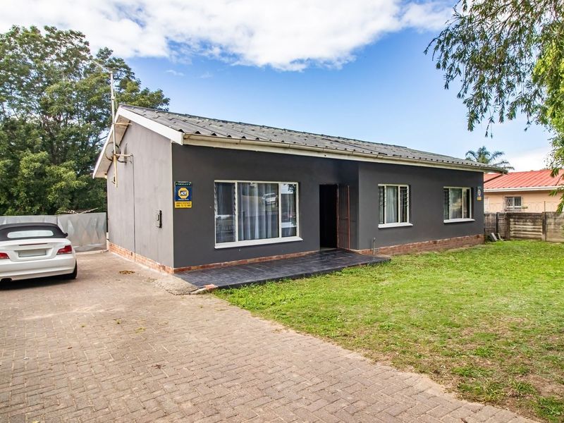 Front view of a detached house featuring a paved driveway, metal roof, and a small tiled entrance porch set within a fenced yard.