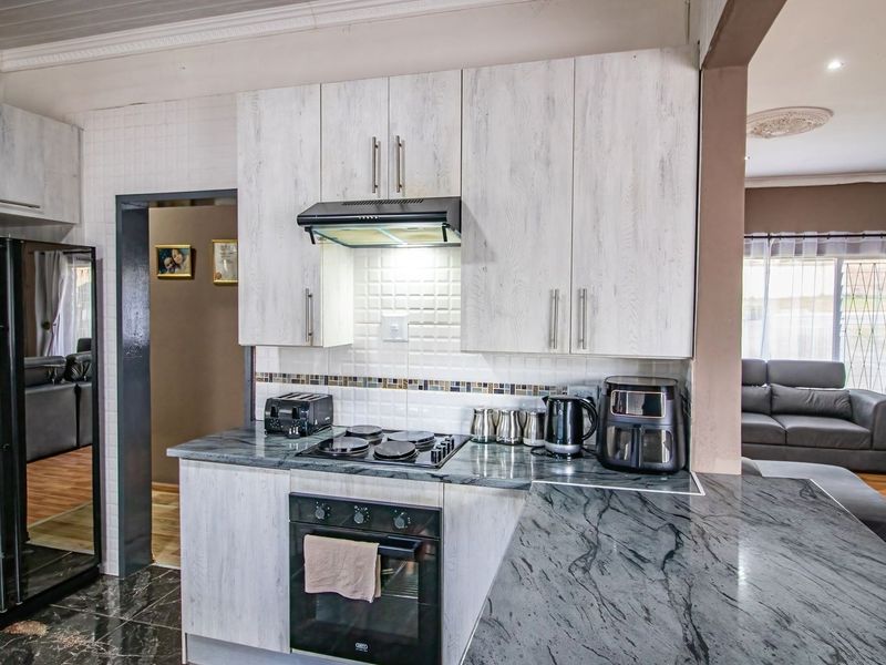 Modern kitchen featuring light wood-grain cabinetry, dark stone countertops, built-in oven, electric hob, and a tiled backsplash, seamlessly connected to the living area.