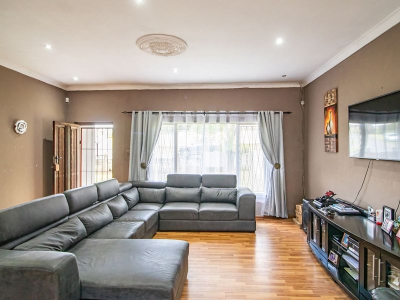 A spacious living room featuring laminate flooring, recessed downlighting, and a decorative ceiling rose, with security features installed on the windows and entrance.