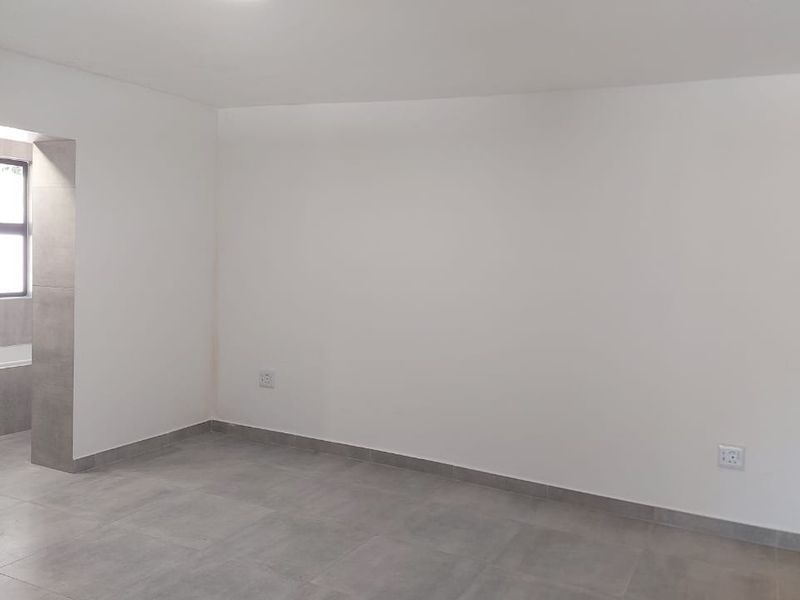 A bright, empty bedroom featuring neutral tiled flooring and clean white walls, with an adjacent tiled bathroom area visible through the doorway.