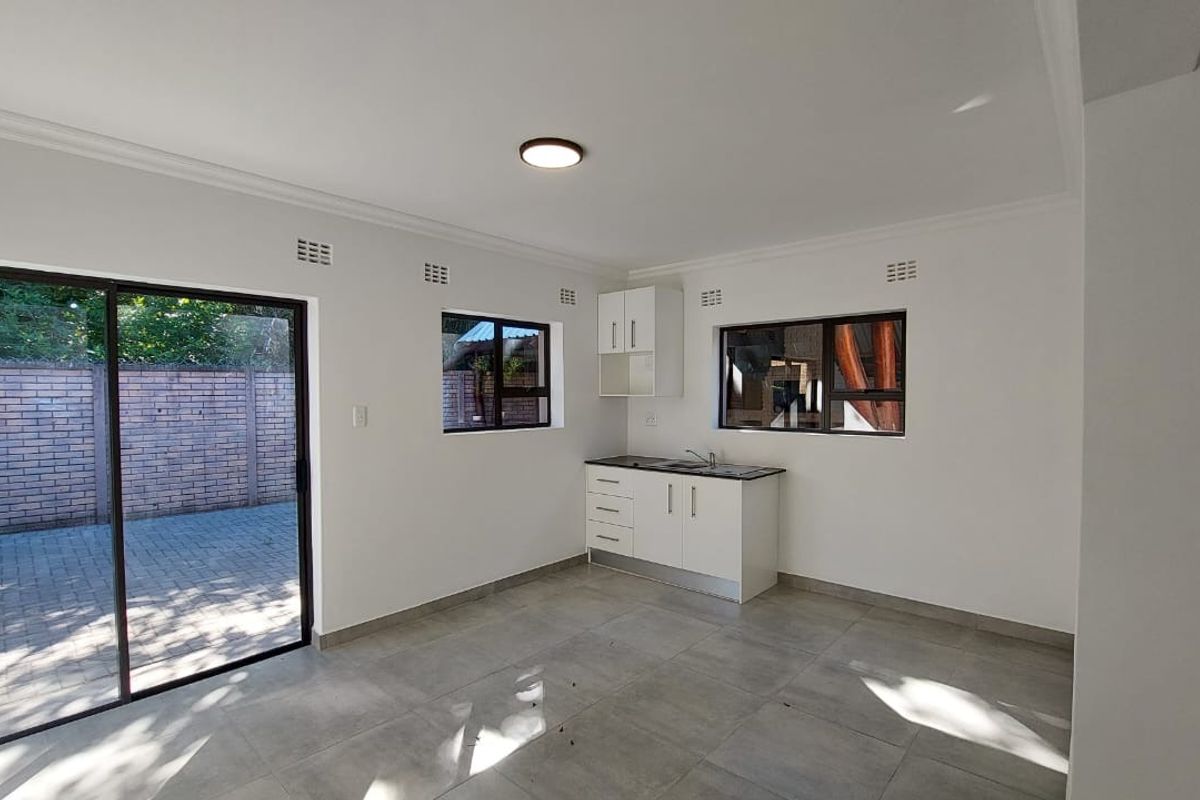 A bright, empty kitchen area featuring modern tiled floors, white cabinetry, a sink, and a sliding glass door leading to an outdoor paved area.