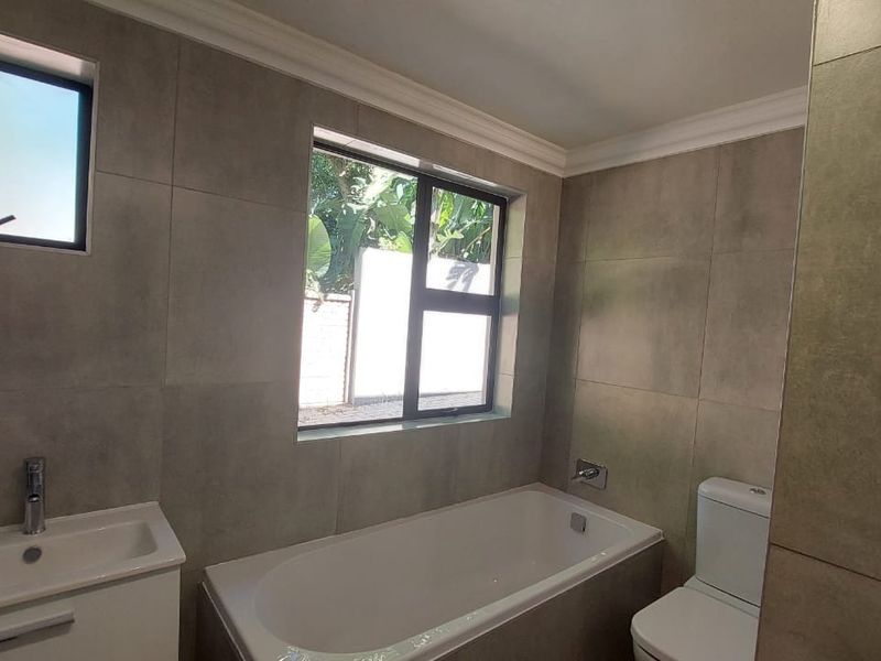 A modern, neutral-toned bathroom featuring a built-in vanity, bathtub, and toilet with floor-to-ceiling tiling and natural light.