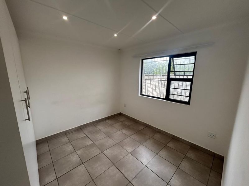 A clean, empty bedroom featuring neutral tiled flooring, built-in cupboards, and a window fitted with security bars.