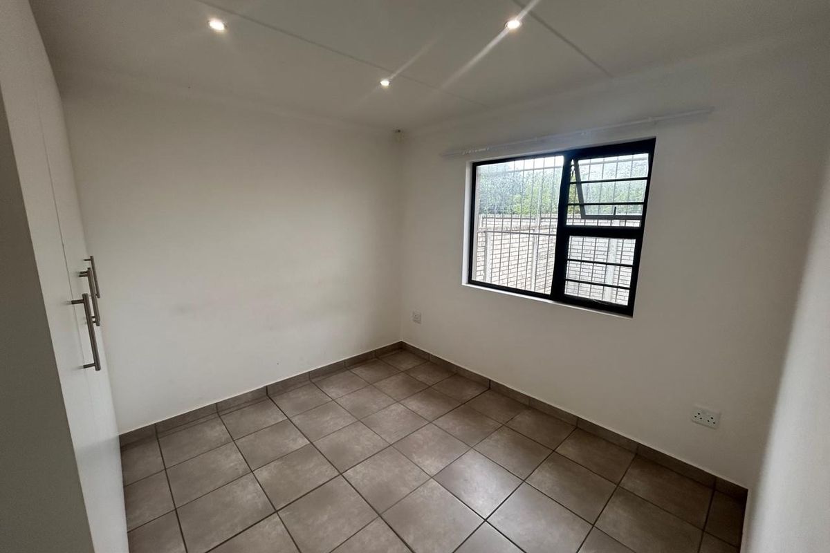 A clean, empty bedroom featuring neutral tiled flooring, built-in cupboards, and a window fitted with security bars.