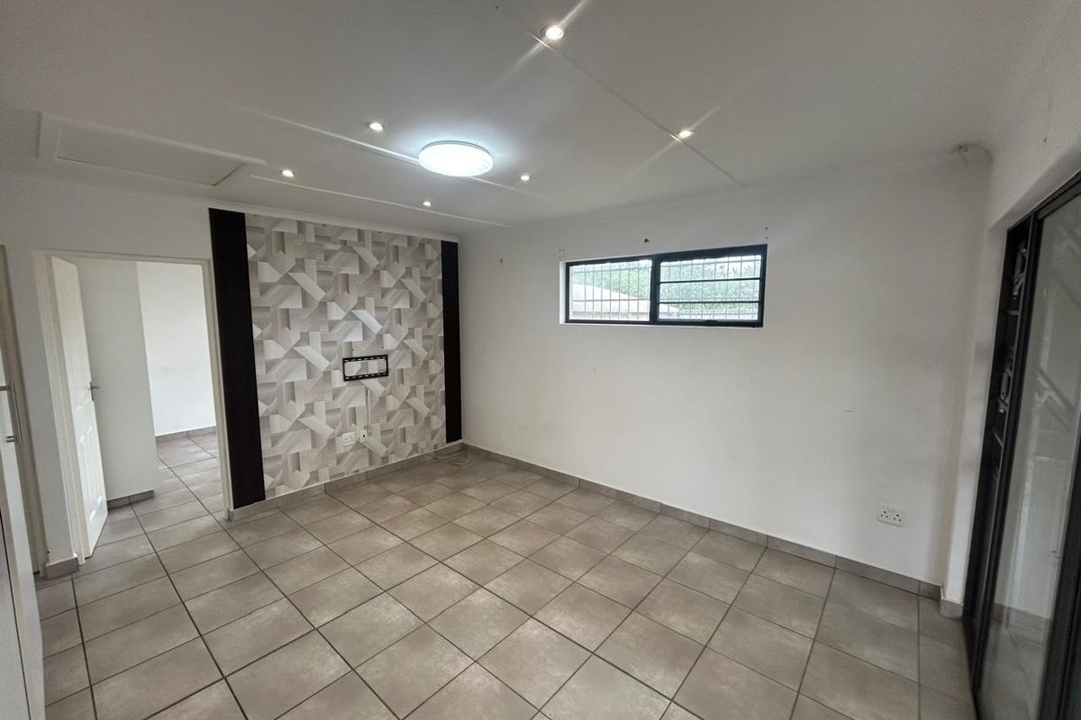 A bright, empty living room featuring neutral floor tiles, a decorative feature wall, recessed lighting, and a sliding door leading to an outdoor area.
