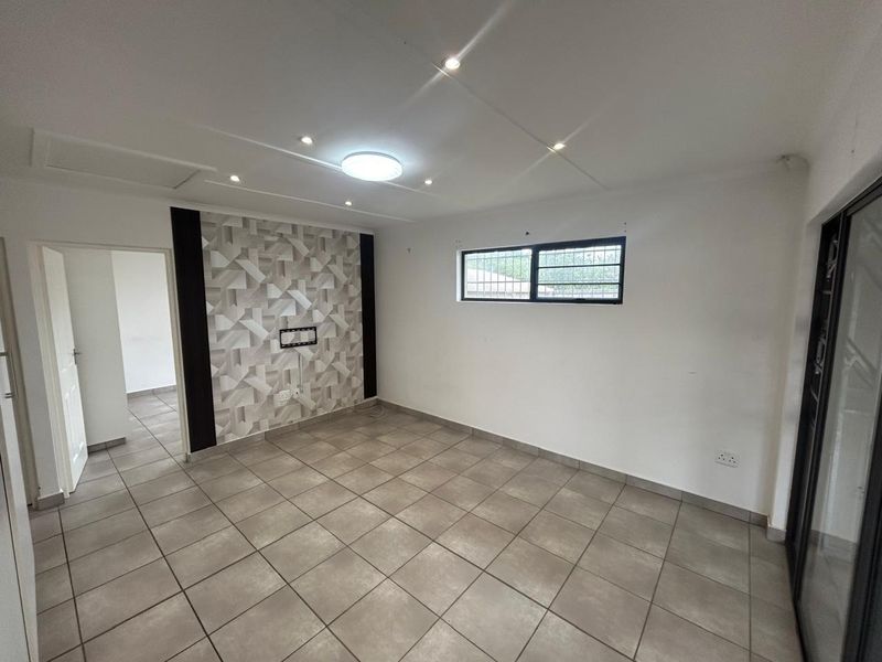 A bright, empty living room featuring neutral floor tiles, a decorative feature wall, recessed lighting, and a sliding door leading to an outdoor area.