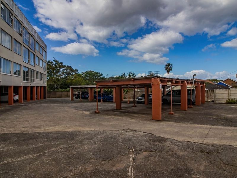 Exterior view of the apartment complex featuring a large concrete parking area with covered bays and multi-story building facade.