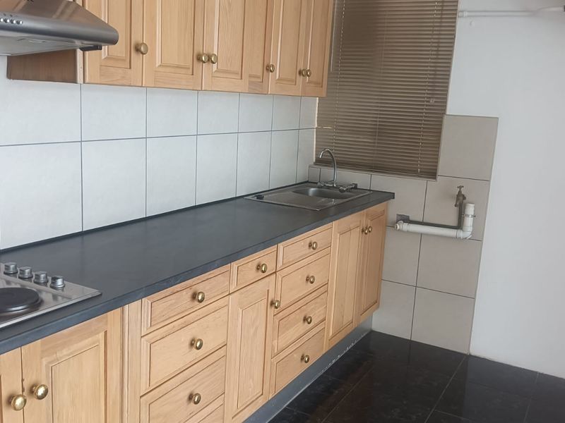 A functional kitchen featuring light wood cabinetry, dark laminate countertops, a built-in electric hob, extractor fan, and tiled backsplash with plumbing for appliances.