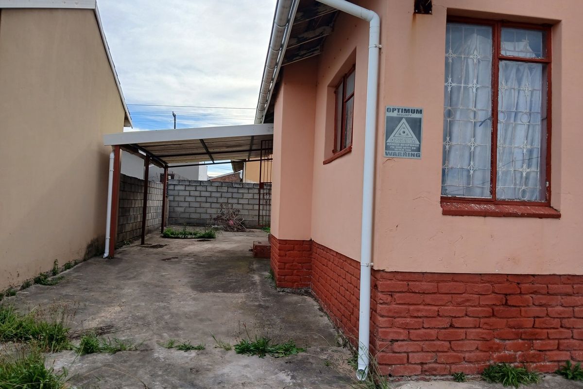 Rear exterior view showing a concrete driveway, a covered carport, and brick boundary walls with visible signs of weathering and maintenance needs.
