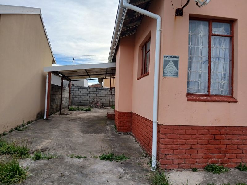 Rear exterior view showing a concrete driveway, a covered carport, and brick boundary walls with visible signs of weathering and maintenance needs.