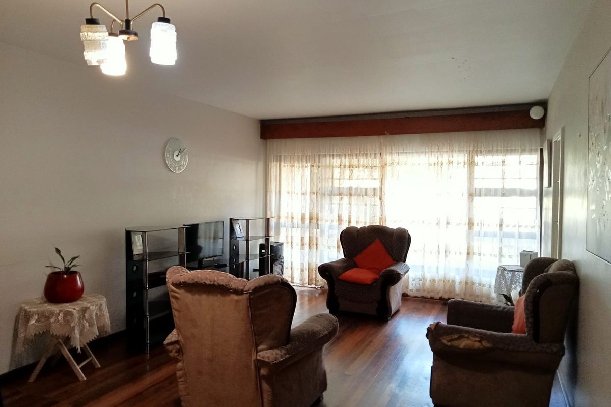 A living room featuring hardwood flooring, a ceiling light fixture, and large windows with curtains, showing signs of wear and dated finishes.