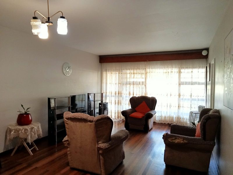 A living room featuring hardwood flooring, a ceiling light fixture, and large windows with curtains, showing signs of wear and dated finishes.
