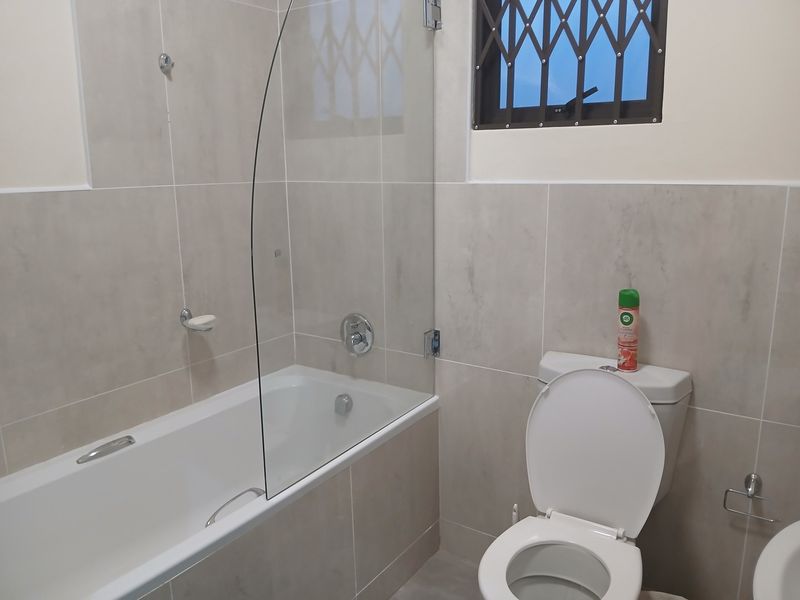 A clean, modern bathroom featuring neutral wall tiles, a bathtub with a glass shower screen, and a secure window with a metal gate.
