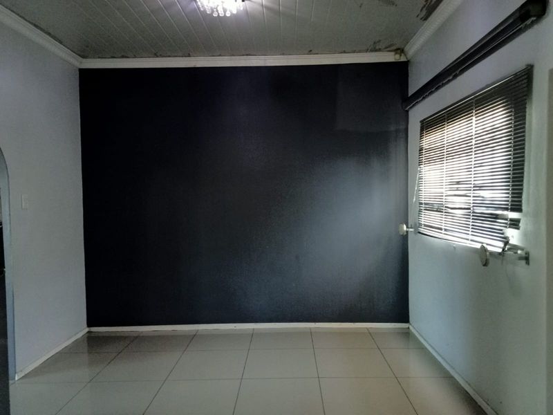 An empty living room featuring tiled flooring, venetian blinds, and ceiling paneling, currently showing signs of ceiling water damage and uneven wall paint.