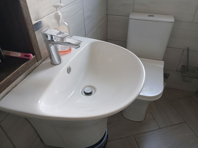A compact bathroom featuring a modern pedestal basin with a chrome mixer tap and a standard toilet, finished with neutral wall and floor tiles.