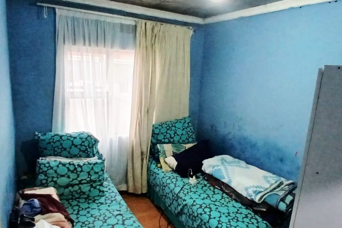 A compact bedroom featuring blue walls and a window with curtain rails, requiring cosmetic updates and wall repairs to improve the living space.