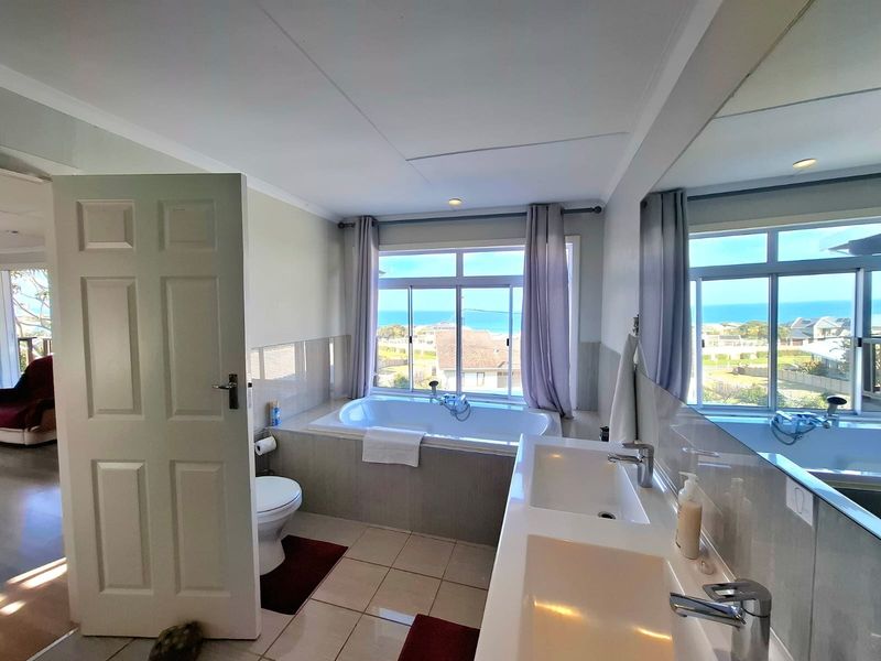 A bright bathroom featuring a built-in bathtub with ocean views, a double vanity with large mirror, and tiled flooring.