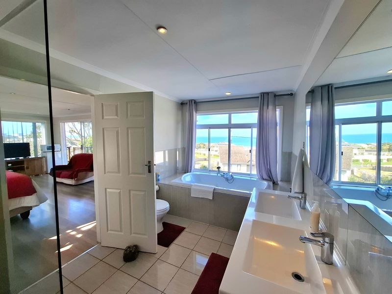 A bright, open-plan bathroom featuring a built-in bathtub, double vanity, and large windows with scenic coastal views.