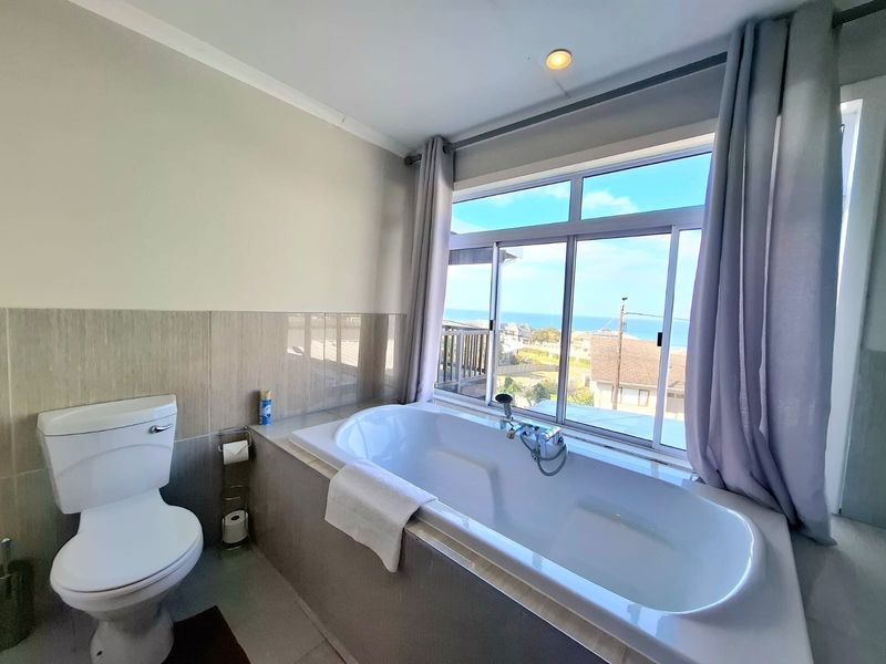 A bright bathroom featuring a built-in bathtub positioned by a large window with ocean views, complemented by a modern toilet and neutral tiling.