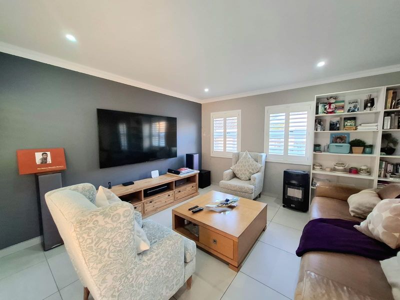 A well-maintained living room featuring tiled flooring, recessed downlights, and plantation shutters, with a dedicated built-in shelving unit.