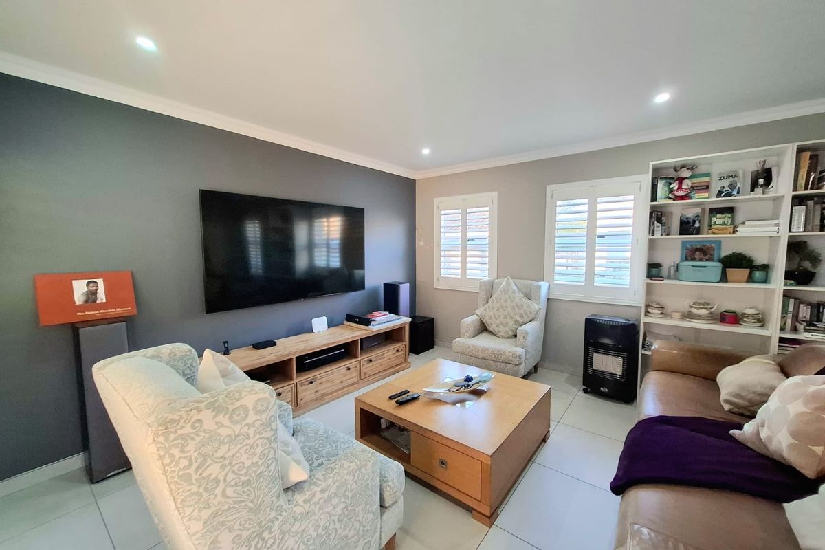 A well-maintained living room featuring tiled flooring, recessed downlights, and plantation shutters, with a dedicated built-in shelving unit.