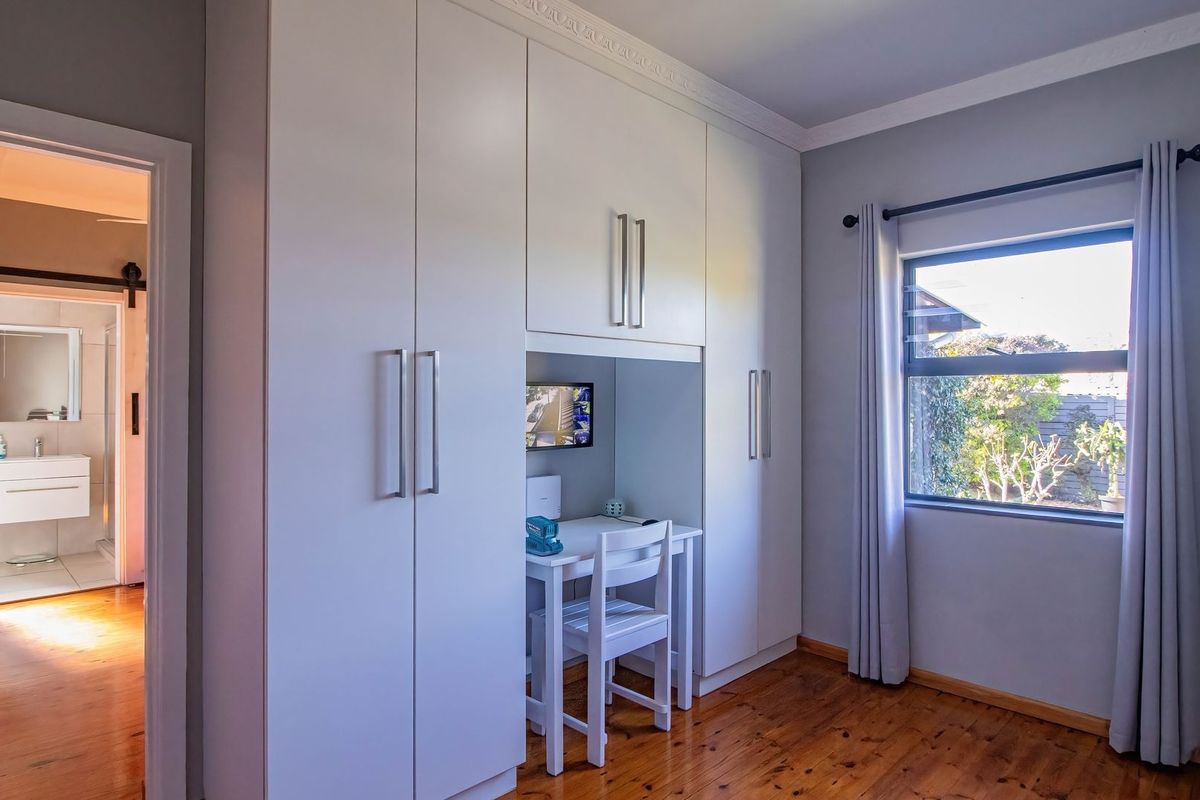 A well-maintained bedroom featuring built-in cupboards with an integrated desk space, polished timber flooring, and a view through an aluminium-framed window.