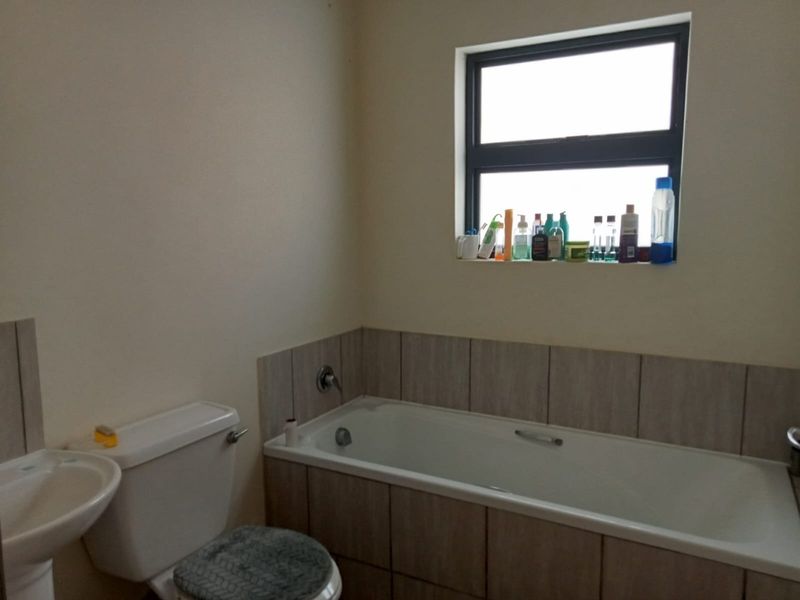 A functional bathroom featuring a bathtub with tiled surround, a pedestal basin, and a toilet, finished with neutral wall tones and an aluminium window.