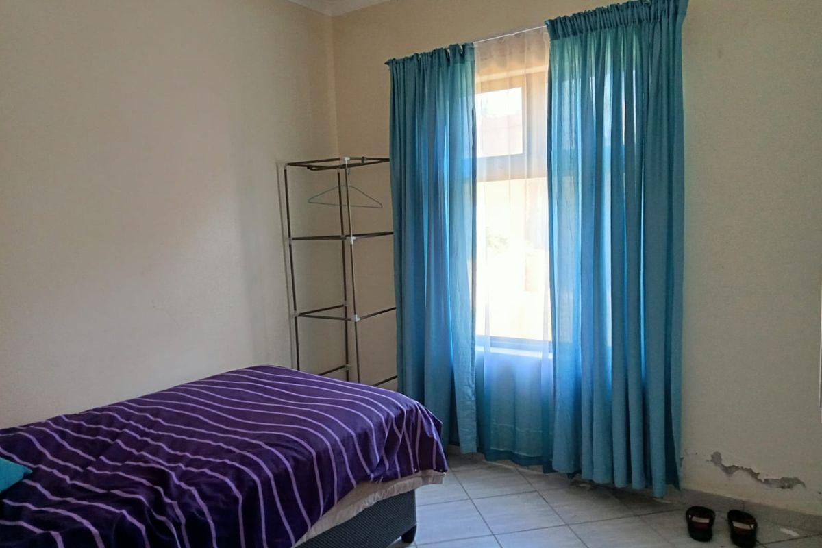 A bedroom featuring tiled flooring and a window with curtains, showing signs of wall surface damage near the base.