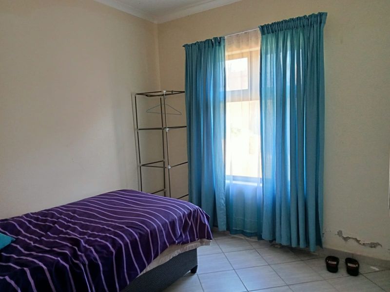 A bedroom featuring tiled flooring and a window with curtains, showing signs of wall surface damage near the base.