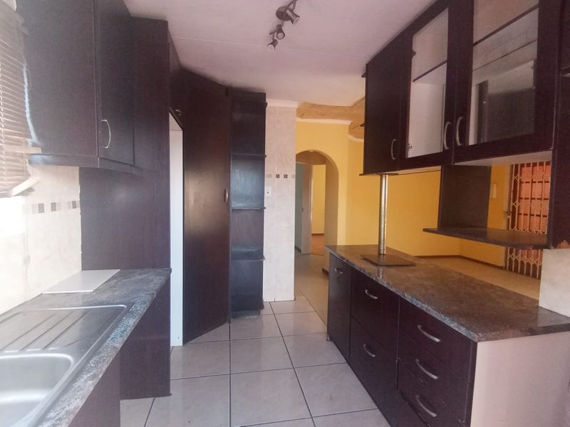A functional kitchen featuring dark built-in cabinetry, laminate countertops, and tiled flooring, leading into an adjacent living area through an arched doorway.