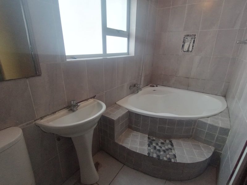 A dated bathroom featuring a pedestal basin, a built-in corner bathtub with tiled surround, and neutral wall tiling throughout.
