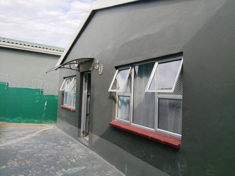 Front exterior view of the property featuring a grey facade, aluminium windows with awnings, and a concrete driveway with perimeter security fencing.